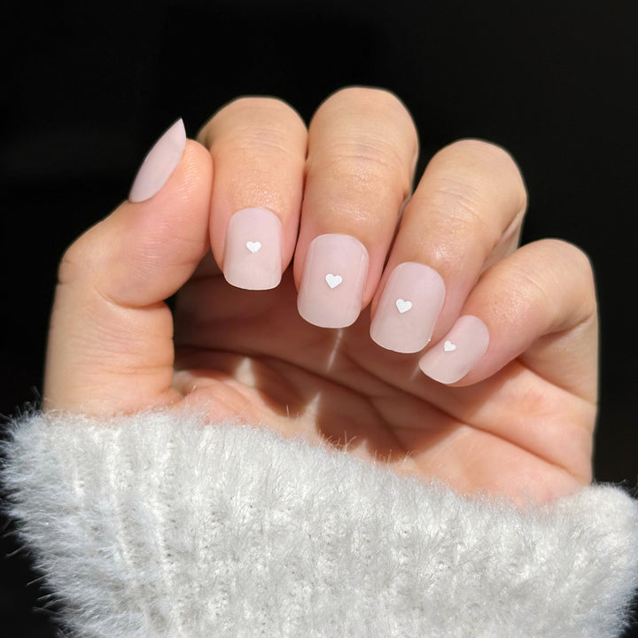 A set of short, square-round press-on nails in a sheer, milky pale pink. Each nail features a small, white 3D heart centered in the middle of the nail bed. The finish is high-gloss, and the overall look is clean, minimalist, and soft.
