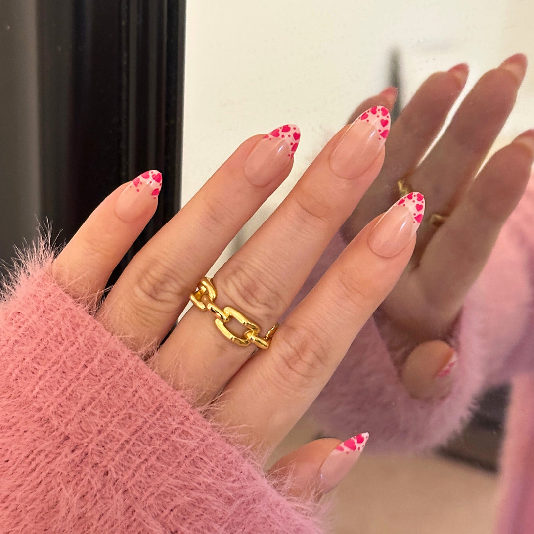 A set of almond-shaped press-on nails with a sheer nude base and bright pink French tips. The tips are decorated with a repeating pattern of small pink hearts and tiny white dots.