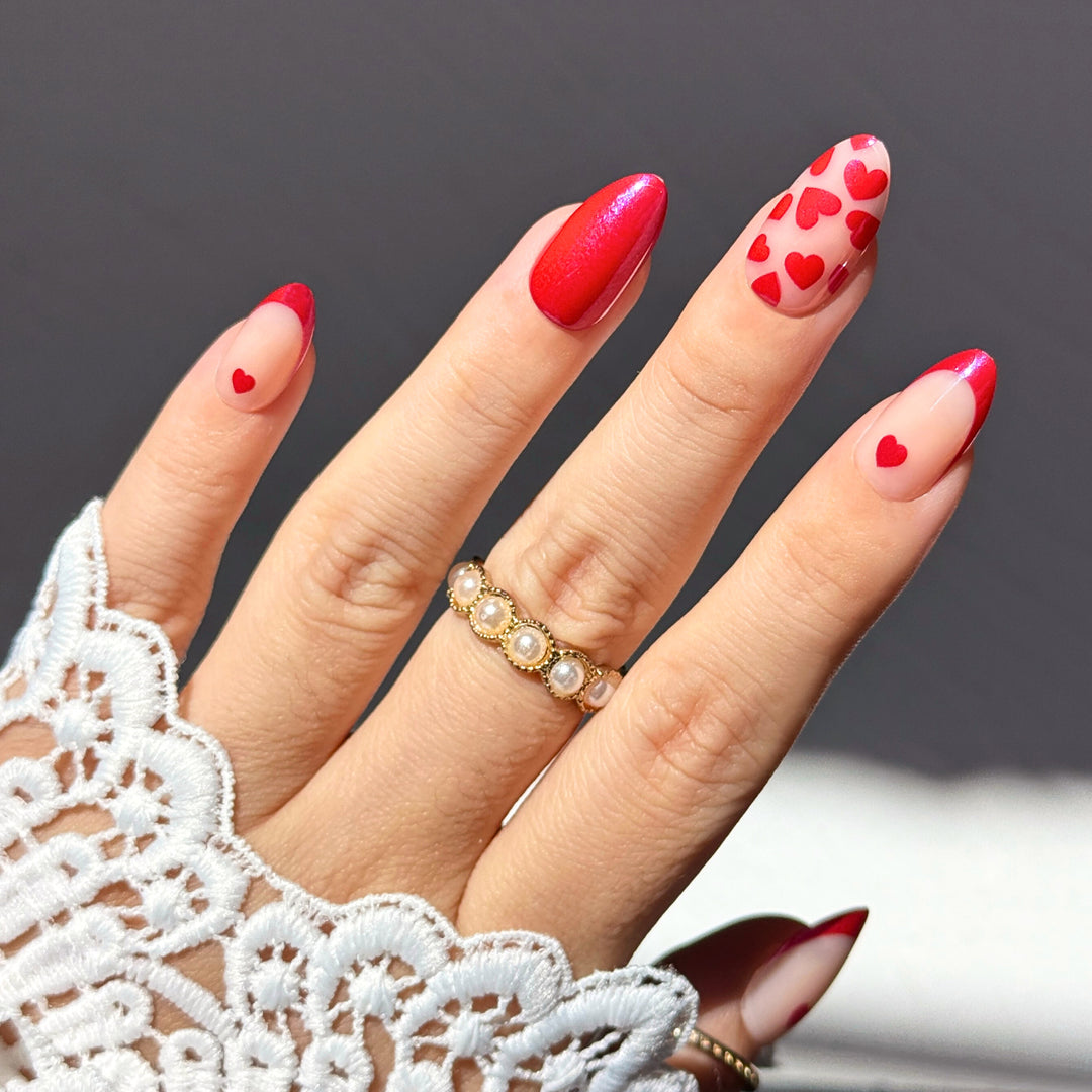 A set of almond press-on nails featuring sheer nude bases with red French tips and small ruby hearts. The collection includes solid shimmering red nails and nails adorned with a repeating heart pattern in a metallic red finish.