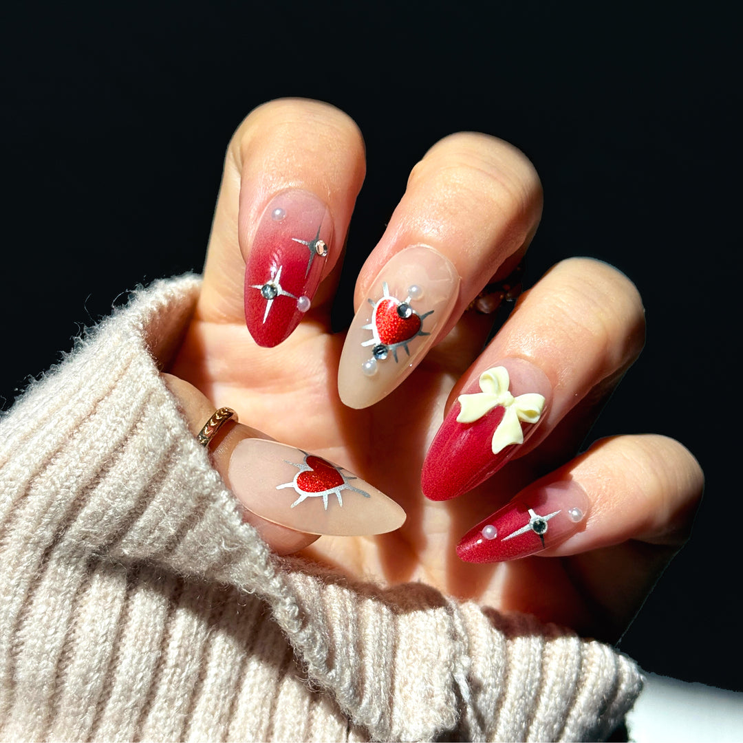 Tapered almond press-on nails with a red aura gradient. The design features silver chrome starbursts, silver-edged red hearts, a large 3D cream bow, and small pearls and rhinestones. The finish is glossy and metallic.