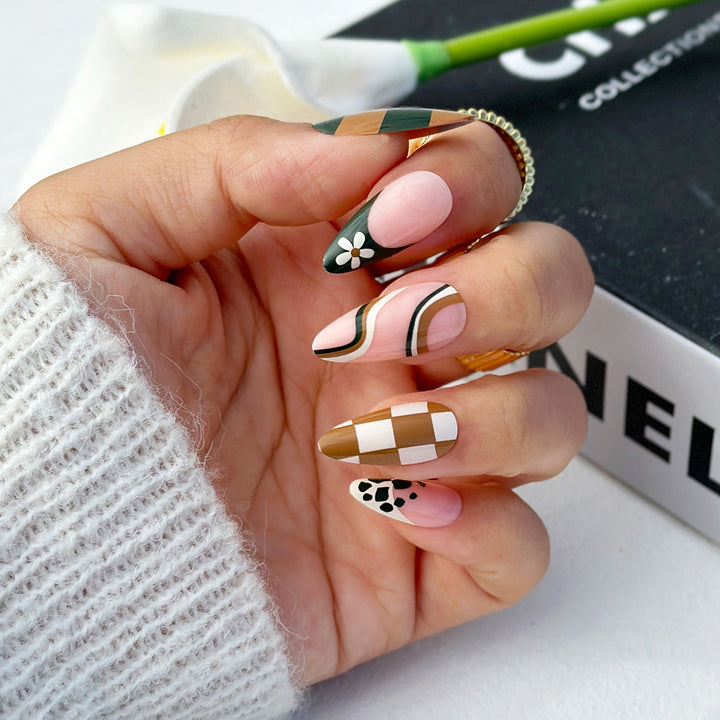 Tapered almond press-on nails featuring a mix of earth-toned designs. Patterns include green and brown checkers, brown and white checkers, wavy stripes, white daisies on green French tips, and black cow print on nude bases.