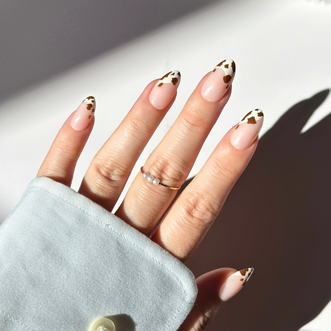 Tapered almond press-on nails with a sheer nude base and dark brown cow print French tips. The nails have a smooth, glossy finish and are shown in a layout and on a hand.