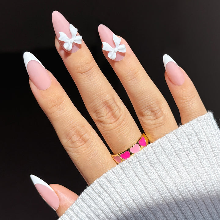 A close-up of a hand showcasing the "First Dance" bridal press-on nail set. The nails feature a matte, soft pink base in a long almond shape, with the thumb and pinky fingers styled in classic white French tips, while the index and middle fingers are accented with large, elegant 3D white bows.