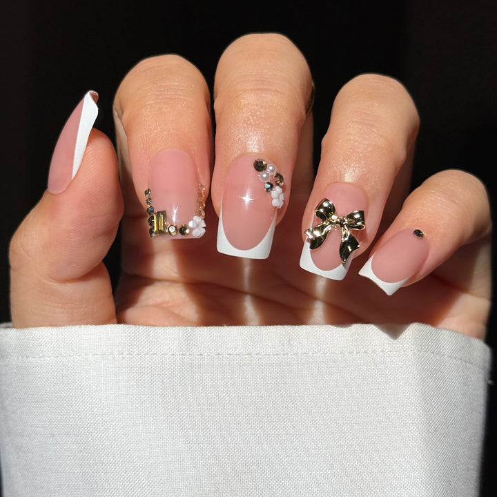 A close-up of a hand showcasing the "Forever Begins" bridal press-on nail set. The nails feature a classic square shape with a glossy nude base and crisp white French tips, dramatically accented with a large 3D metallic gold bow on the ring finger, and an opulent mix of gold crystals, rhinestones, pearls, and mini white flowers decorating the cuticles and side walls of the index and middle fingers.