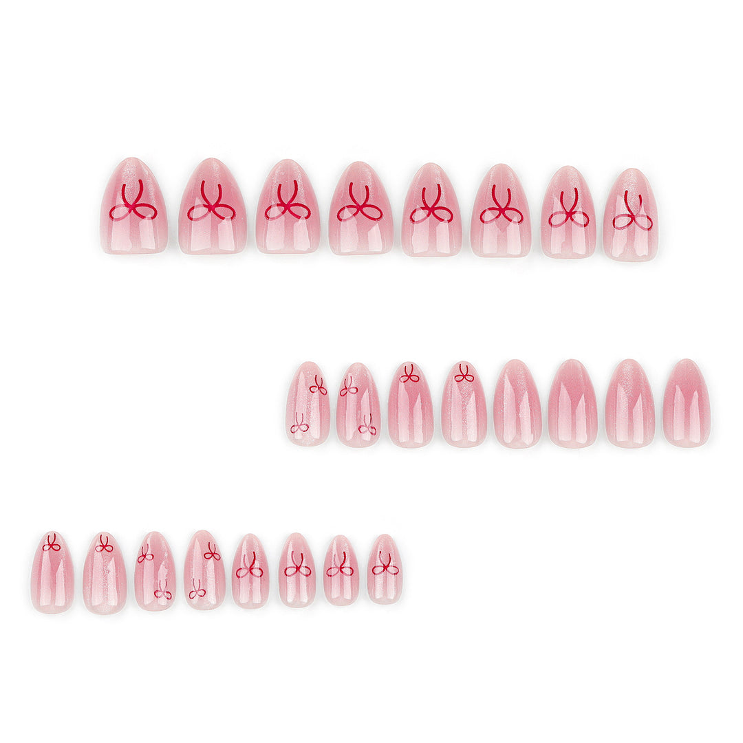 Almond-shaped press-on nails with a sheer blush pink base and subtle pearlescent shimmer, featuring small glossy red ribbon bow designs centered on each nail. The nails are displayed in multiple sizes on a clean white background, showing a smooth, high-gloss finish.