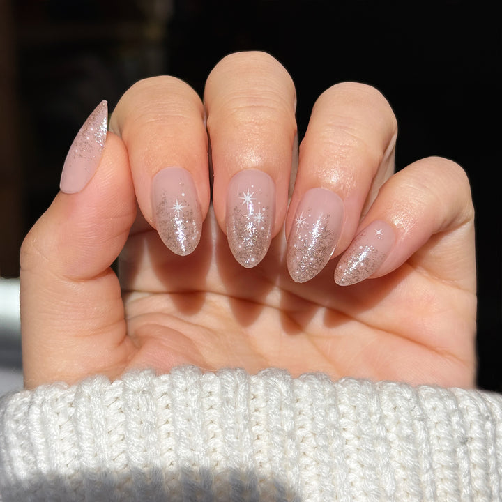 Soft nude almond-shaped press-on nails with a sheer base, silver glitter fade at the tips, and delicate white star accents, shown on a hand against a dark background.