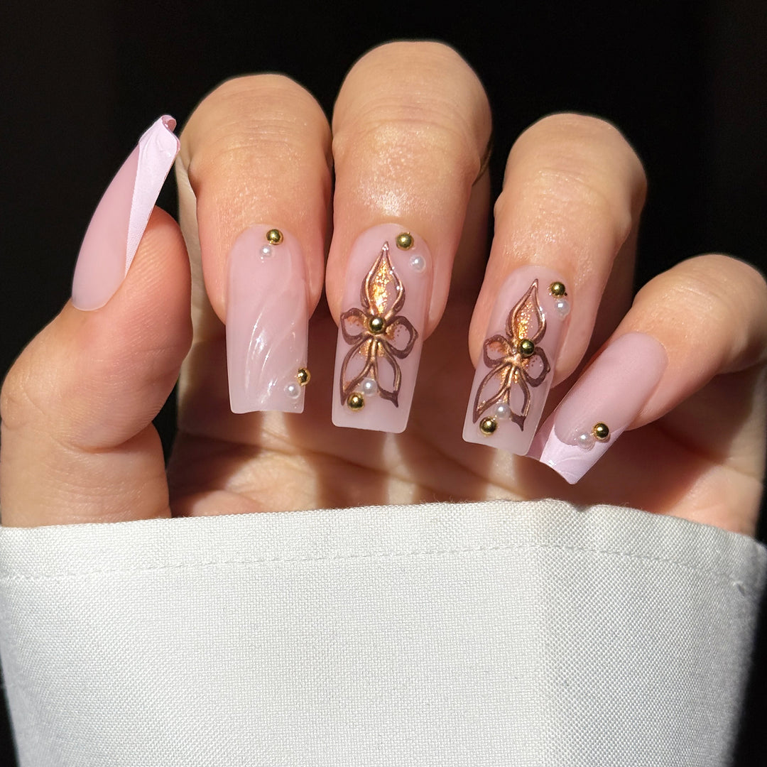 A close-up image showcasing the "Peony Promise" bridal press-on nail set. The long, square-shaped nails feature a soft, semi-sheer milky pink base decorated with intricate hand-painted metallic rose gold and bronze floral designs on the middle and ring fingers, textured 3D swirl patterns on the index finger and French tip, and a delicate placement of tiny pearls and reflective gold studs near the cuticles.