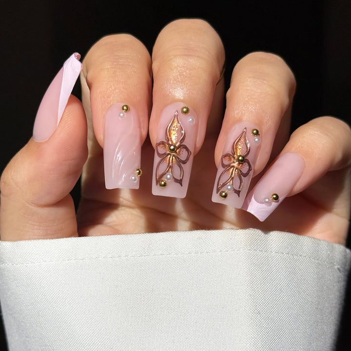 A close-up image showcasing the "Peony Promise" bridal press-on nail set. The long, square-shaped nails feature a soft, semi-sheer milky pink base decorated with intricate hand-painted metallic rose gold and bronze floral designs on the middle and ring fingers, textured 3D swirl patterns on the index finger and French tip, and a delicate placement of tiny pearls and reflective gold studs near the cuticles.