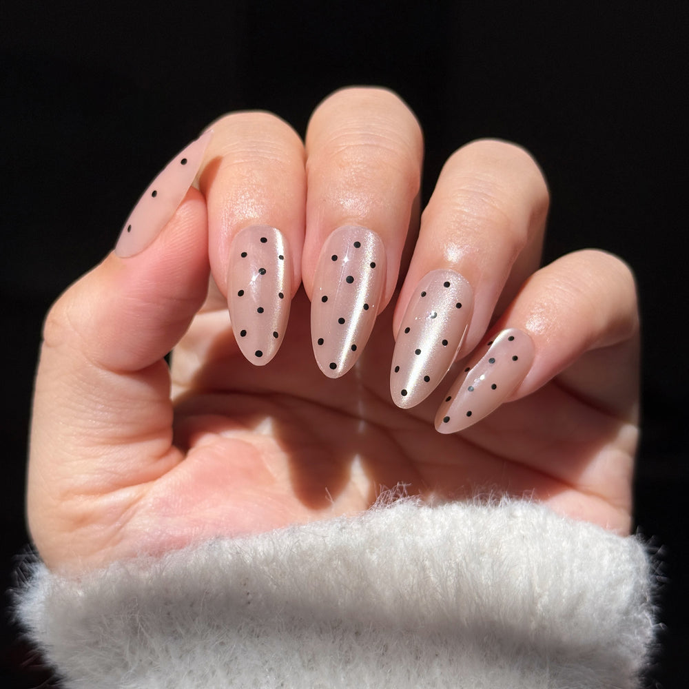 Almond-shaped press-on nails featuring a shimmering iridescent nude base and a minimalist black polka dot pattern.