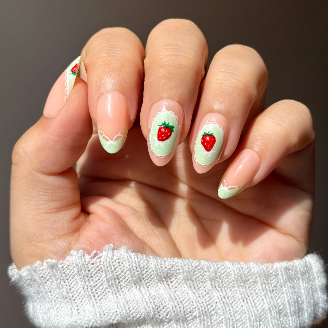 Light pink press-on nails with light green French tips, white lace patterns, and small red strawberry nail art. The middle nail features a single strawberry inside a white lace frame.