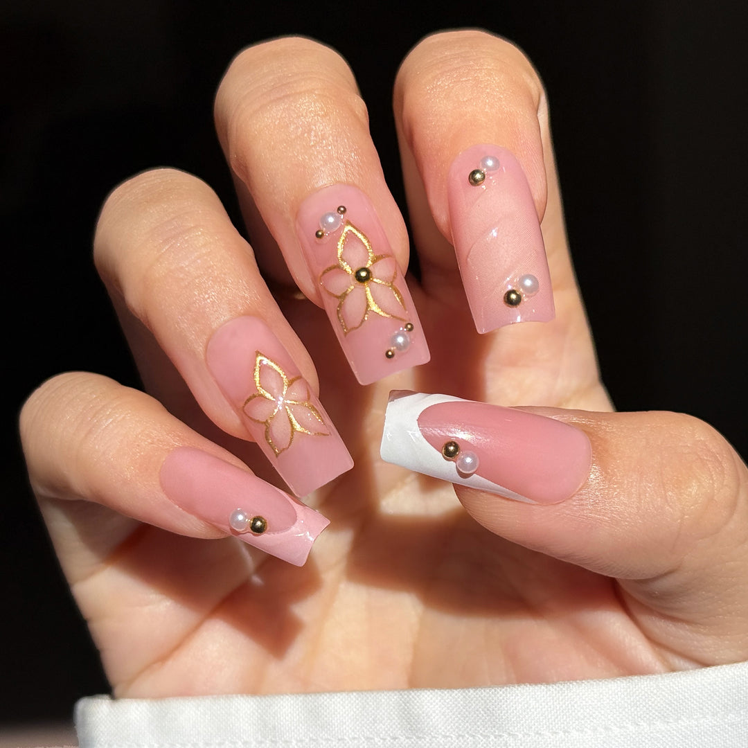 A close-up image displaying the "Sweetest Sayings" bridal press-on nail set. The long, square nails feature a soft, translucent jelly pink base, beautifully detailed with intricate hand-painted metallic gold outlines of five-petal blossoms on the index and middle fingers. Subtle textured 3D swirl patterns embellish the ring finger and the elegant white French tips, while clusters of miniature white pearls and shiny gold studs accent the cuticles and design borders.