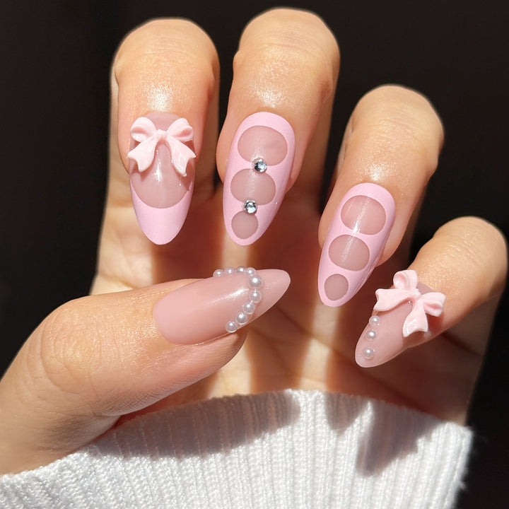 A close-up of a hand showcasing the "Tie The Knot" coquette bridal press-on nail set. The nails feature a mix of glossy nude and soft pastel pink bases, detailed with 3D pink bows on the index and pinky fingers, negative-space circle cutouts with rhinestones on the middle and ring fingers, and an elegant curve of pearls on the thumb.