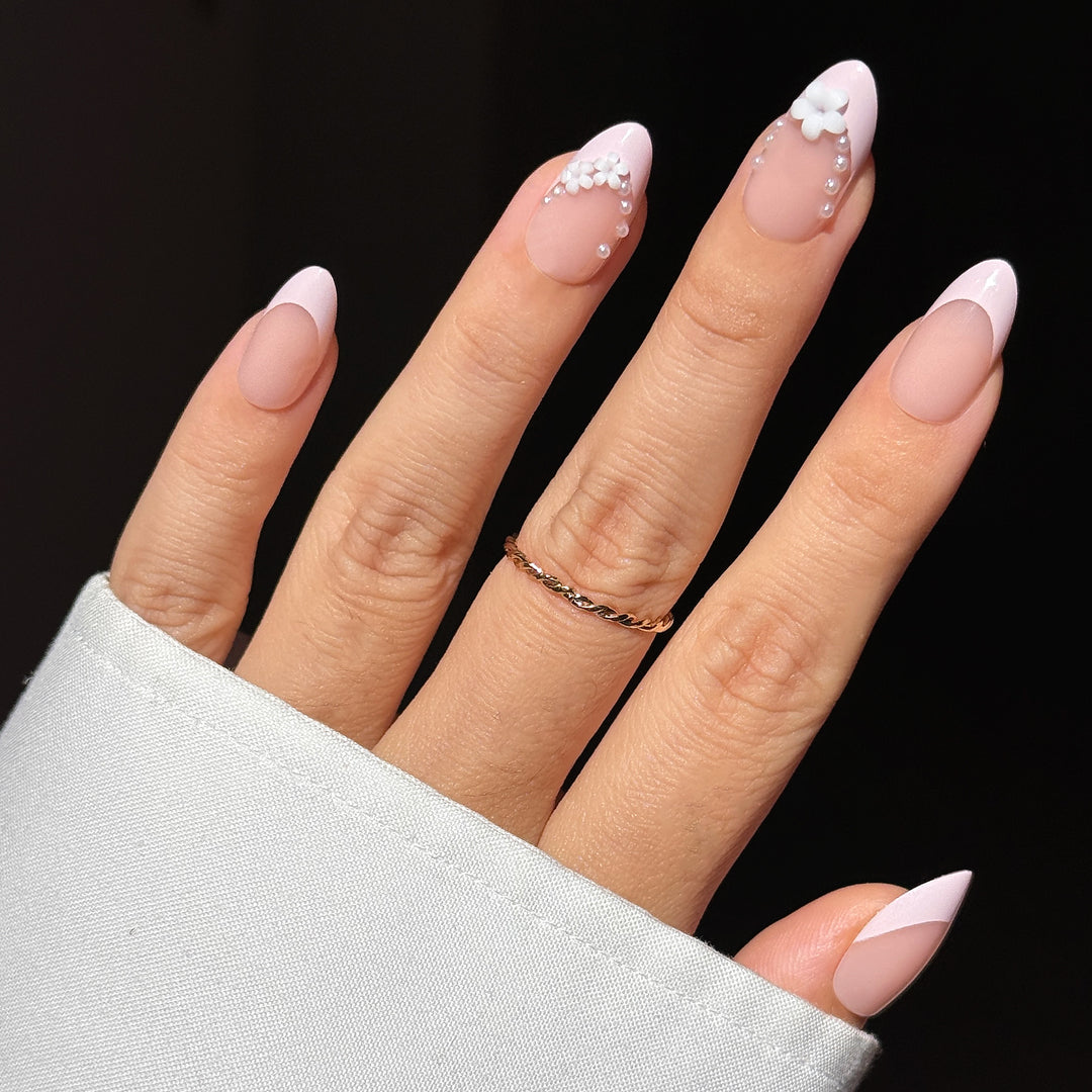 A close-up of a hand showcasing the "To Have & Hold" bridal press-on nail set. The medium almond-shaped nails feature a soft matte nude base with elegant, glossy pale pink French tips, beautifully accented with white 3D floral blossoms and delicate pearl framing along the French smile lines on the index and middle fingers.