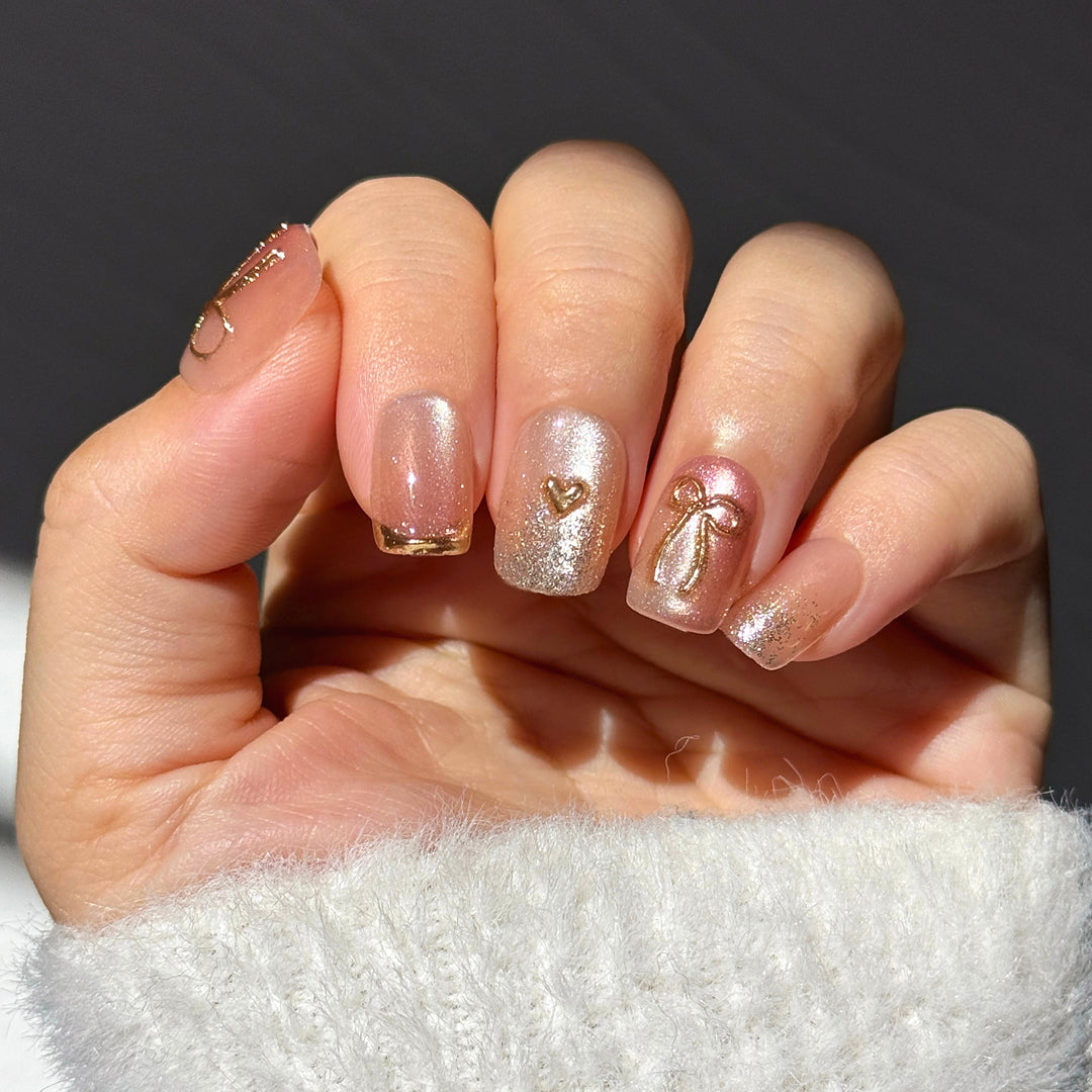Short square press-on nails with a shimmering rose-pink aura base. The set features 3D gold metallic bow and heart charms, gold glitter gradients, and thin gold metallic tips. The design has a high-gloss finish and a luxury aesthetic.